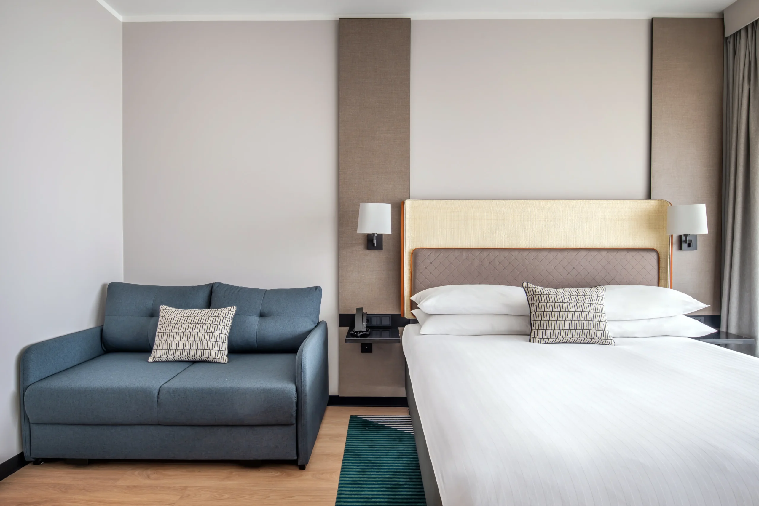 Wykończenia wnętrz nowoczesnego pokoju z dużym łóżkiem, tapicerowanym zagłówkiem, sofą i kinkietami