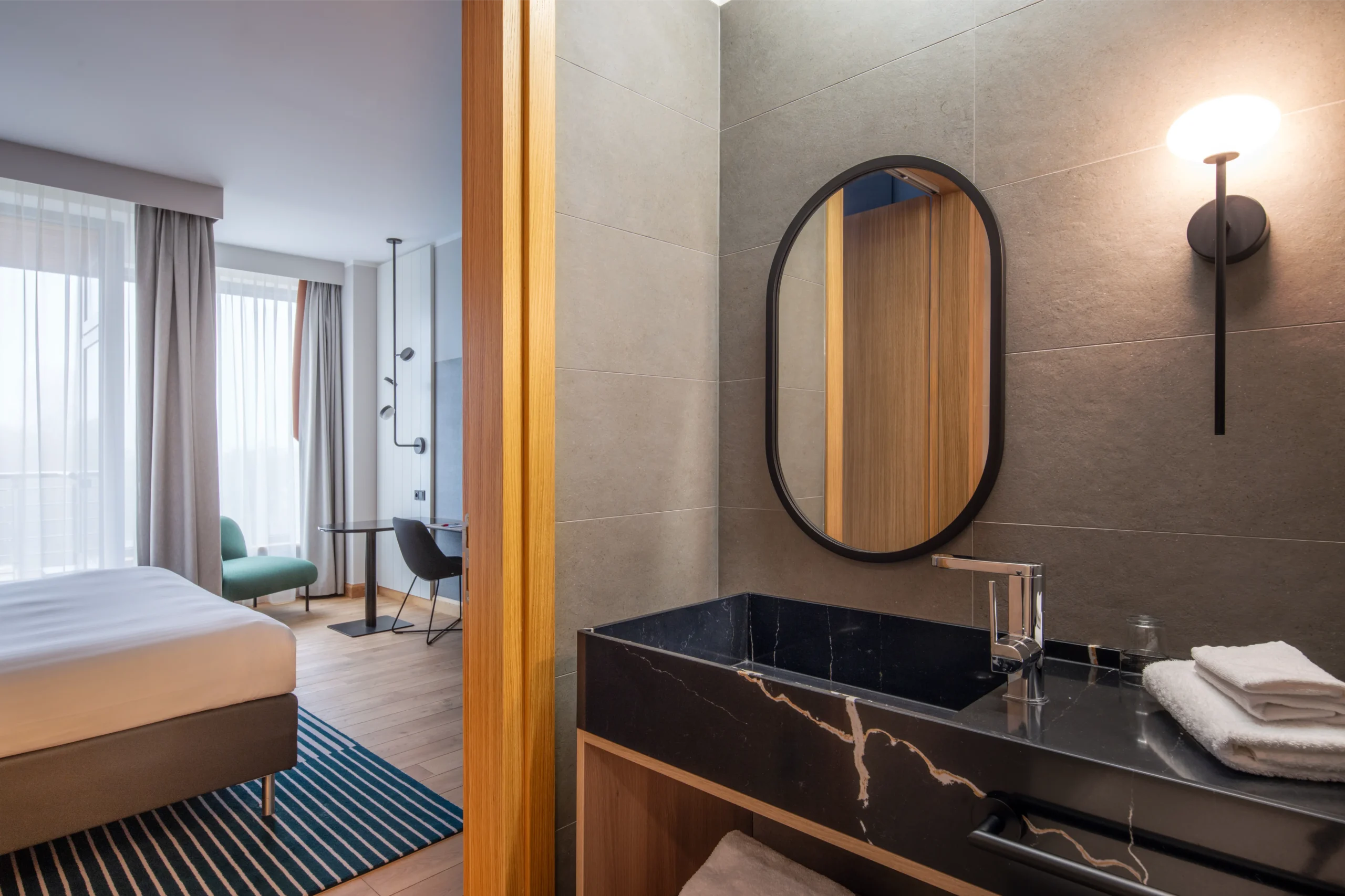 Fit-out wnętrza hotelowego: łazienka z czarnym marmurowym blatem, owalnym lustrem i przyległa sypialnia