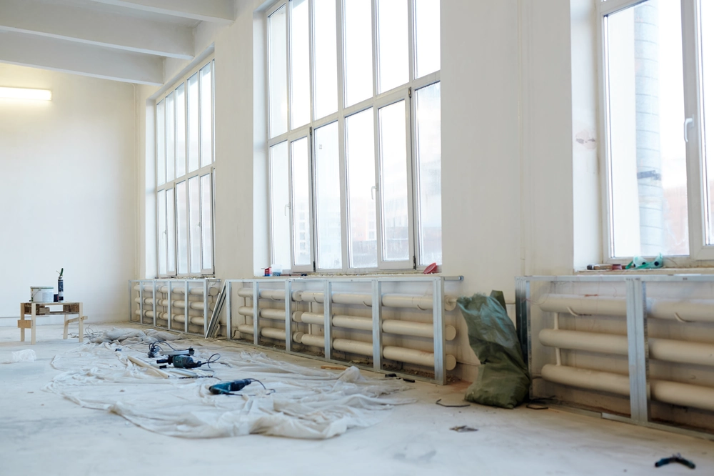 Wnętrze biura w trakcie remontu z dużymi oknami, odsłoniętymi grzejnikami, narzędziami i folią ochronną na podłodze