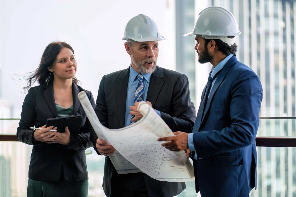 Trzech specjalistów w kaskach omawia plany budowlane na tle biurowców (zarządzanie projektami budowlanymi)